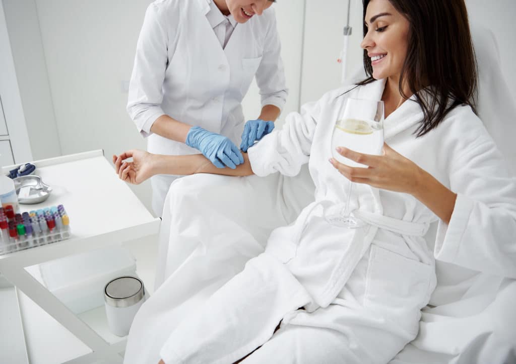 Smiling Woman in Bathrobe sitting in Armchair and Receiving IV infusion with Holding Glass on Hands | Skinlogicx in Queens, NY